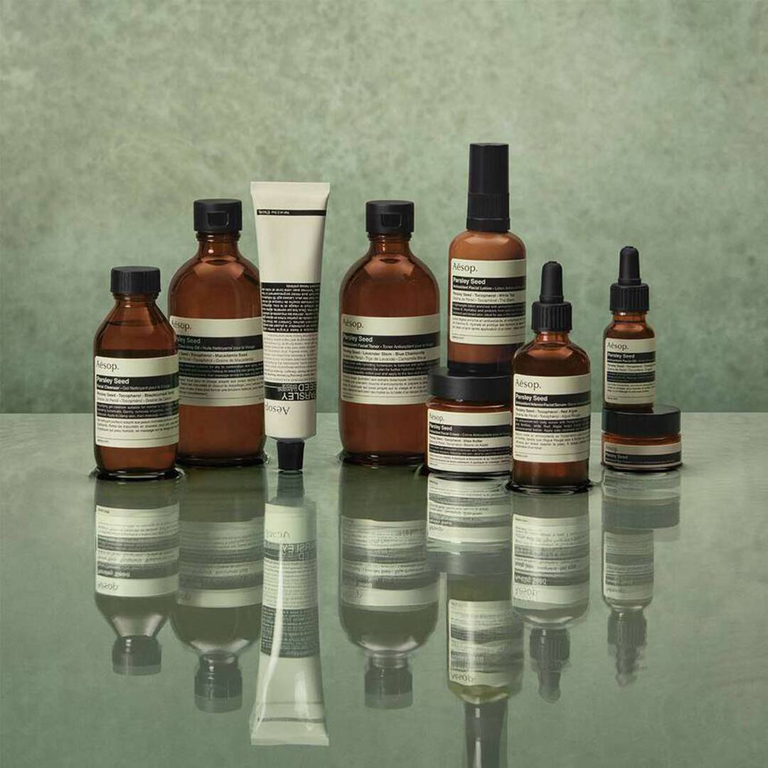 Amber Aesop skincare bottles, jars, and dropper serums arranged on a reflective surface against a muted green background.
