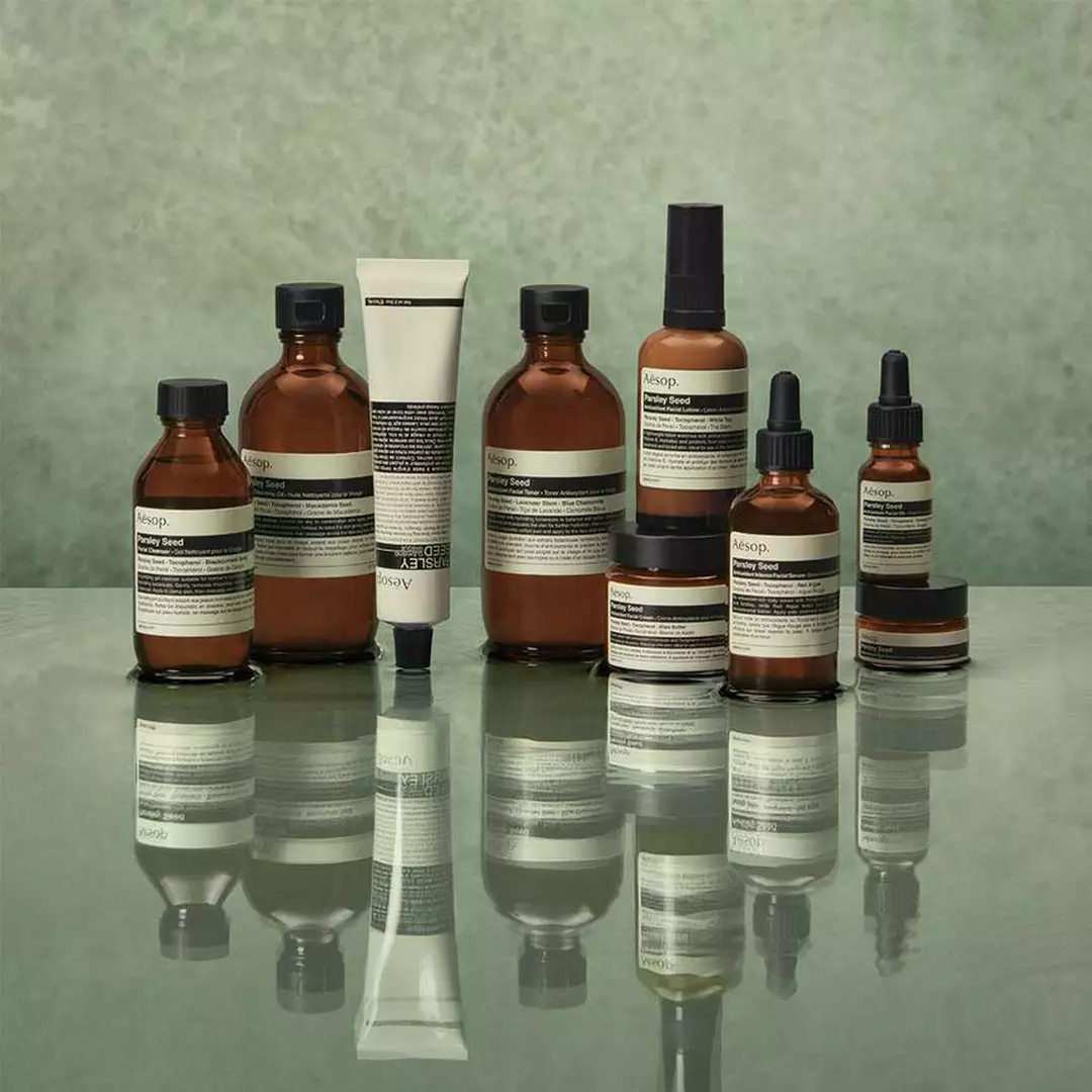 Amber Aesop skincare bottles, jars, and dropper serums arranged on a reflective surface against a muted green background.