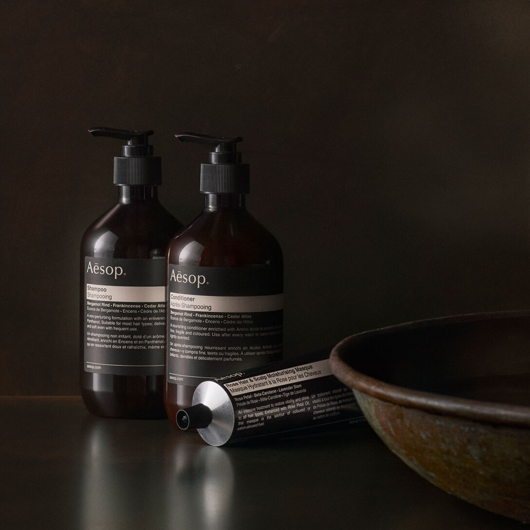 Aesop shampoo, conditioner, and hair masque in a dark, minimalist setting next to a ceramic bowl.