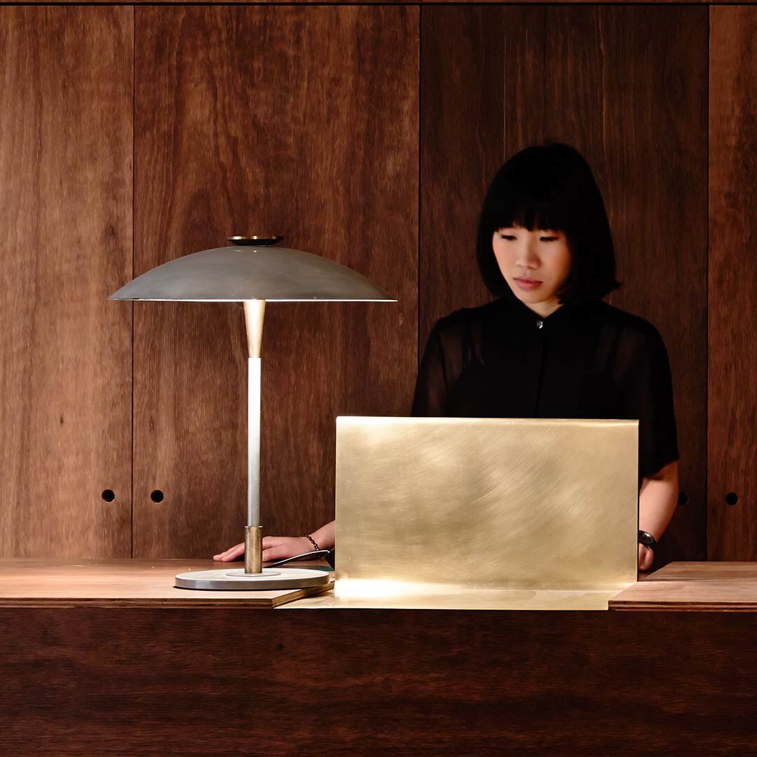 A photograph of a woman with short black hair and bangs, wearing a black shirt, looking downward behind a minimalist reception desk. On the desk sits a sleek brushed-brass laptop stand or screen and a modern metal desk lamp with a mushroom-style shade casting a warm glow. The background consists of vertical, rich dark-brown wood panels.