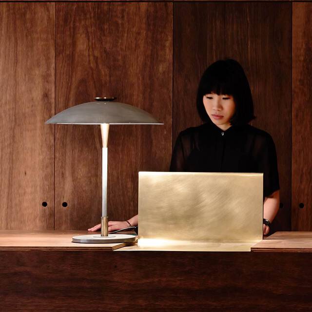 A photograph of a woman with short black hair and bangs, wearing a black shirt, looking downward behind a minimalist reception desk. On the desk sits a sleek brushed-brass laptop stand or screen and a modern metal desk lamp with a mushroom-style shade casting a warm glow. The background consists of vertical, rich dark-brown wood panels.