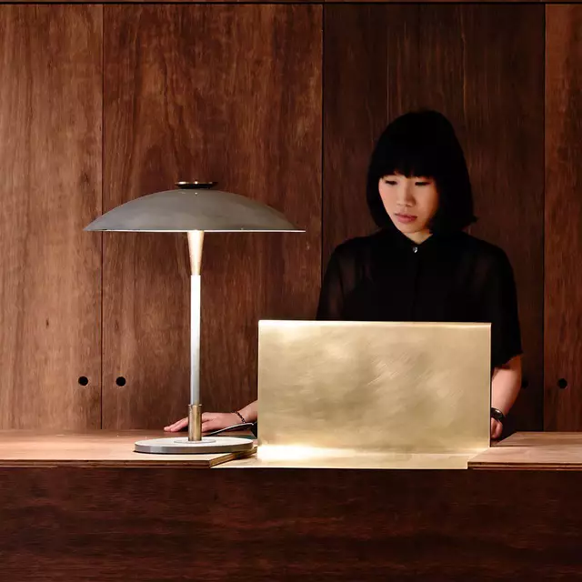 A photograph of a woman with short black hair and bangs, wearing a black shirt, looking downward behind a minimalist reception desk. On the desk sits a sleek brushed-brass laptop stand or screen and a modern metal desk lamp with a mushroom-style shade casting a warm glow. The background consists of vertical, rich dark-brown wood panels.