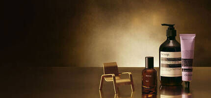 An Aesop perfume bottle, a miniature gold chair, a delicate gold lamp, an Aesop pump bottle, and an Aesop hand cream tube are artfully arranged on a reflective surface against a gradient dark to golden-brown background.