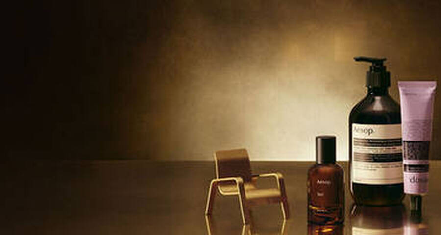 An Aesop perfume bottle, a miniature gold chair, a delicate gold lamp, an Aesop pump bottle, and an Aesop hand cream tube are artfully arranged on a reflective surface against a gradient dark to golden-brown background.