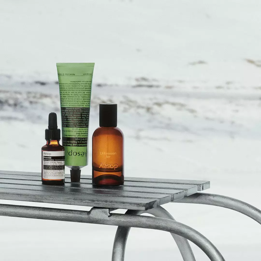 An amber dropper bottle, a tall green Aesop tube and an amber bottle with a black cap stand on a metal sledge in snow.
