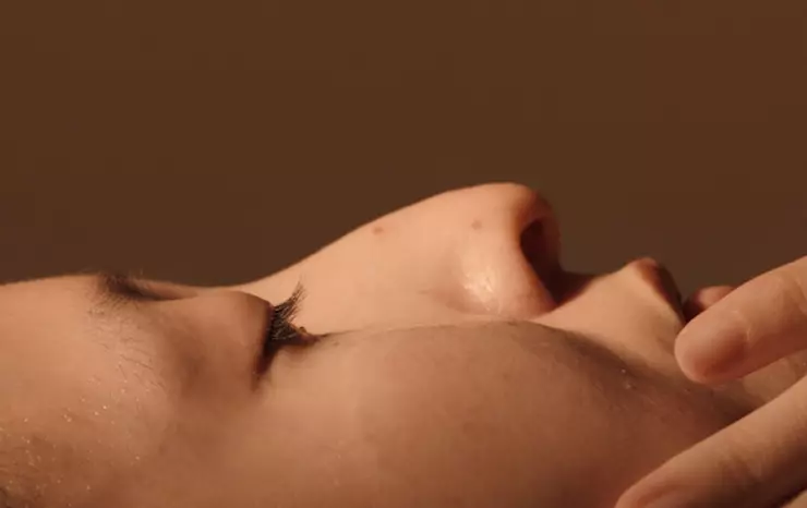 A close-up, side-profile view of a person lying down with their eyes closed. The warm, soft-focus lighting highlights the contours of their nose, long eyelashes, cheek, and upper lip. A hand is shown gently touching the lower part of their face near the chin, set against a solid, warm brown background.