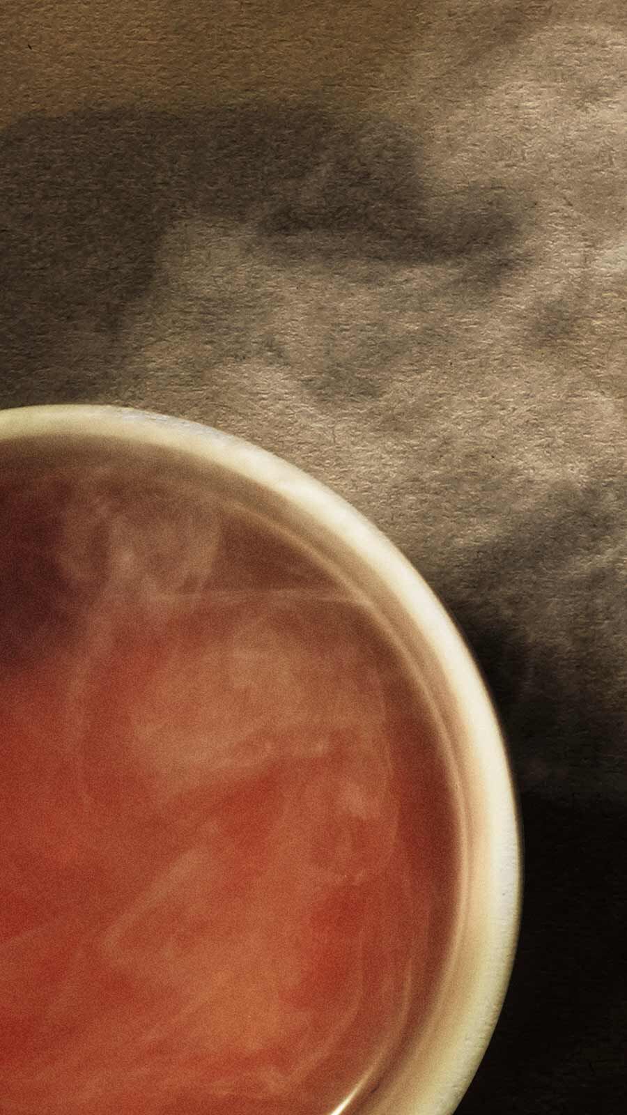 Small ceramic bowl filled with warm red-orange liquid with wisps of steam against a soft grey background.