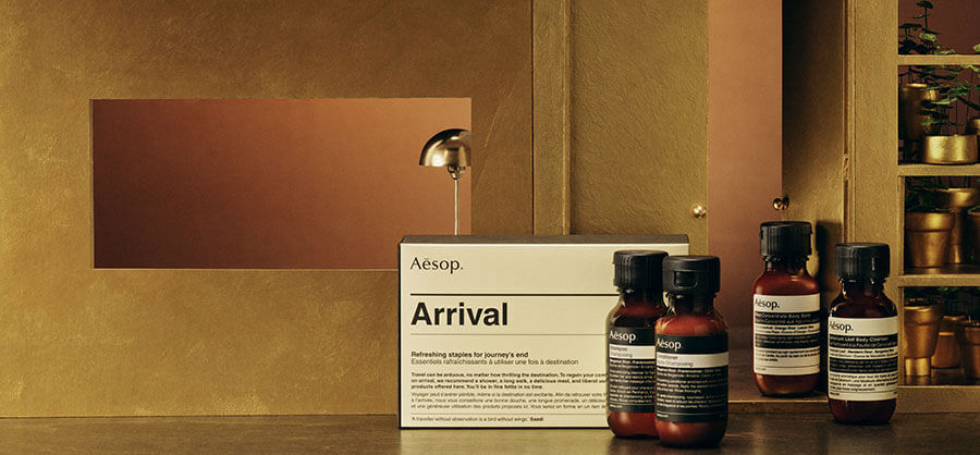 An "Aesop Arrival" kit box and several individual Aesop product bottles are displayed on a dark wooden surface in front of a golden background with shelves holding potted plants.