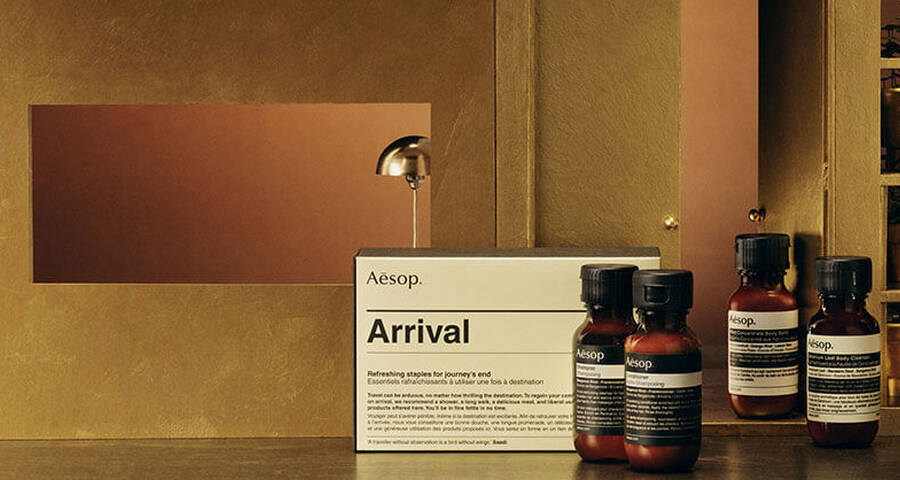 An "Aesop Arrival" kit box and several individual Aesop product bottles are displayed on a dark wooden surface in front of a golden background with shelves holding potted plants.