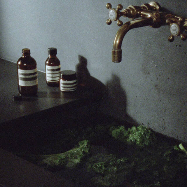 A moody, dark stone sink featuring an antique brass faucet, with green leafy vegetables in the basin and three amber skincare bottles resting on the adjacent ledge.