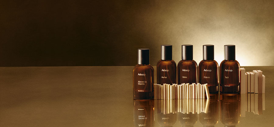 Six amber-colored Aesop perfume bottles with black caps are lined up on a reflective, golden-brown surface, with small rectangular blocks in front of some.