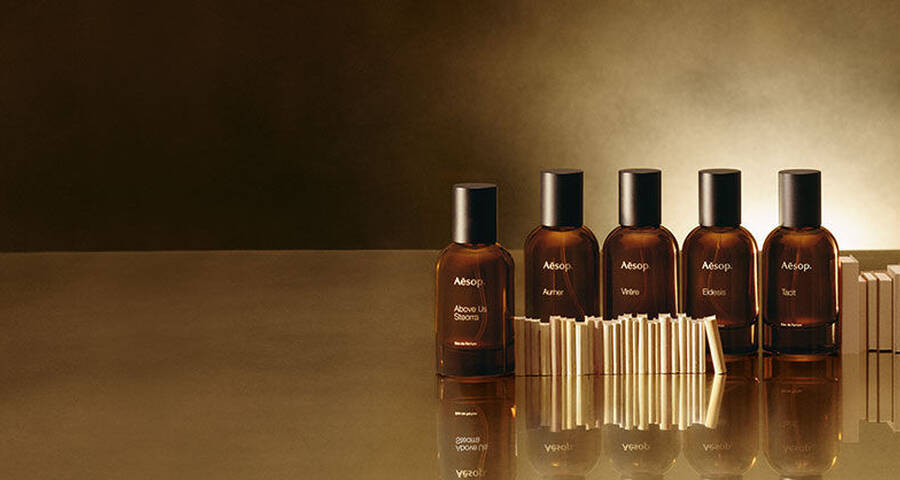 Six amber-colored Aesop perfume bottles with black caps are lined up on a reflective, golden-brown surface, with small rectangular blocks in front of some.