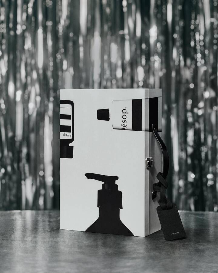 Upright silver Aesop case decorated with black bottle silhouettes, with a black luggage tag resting on a table against silver tinsel.