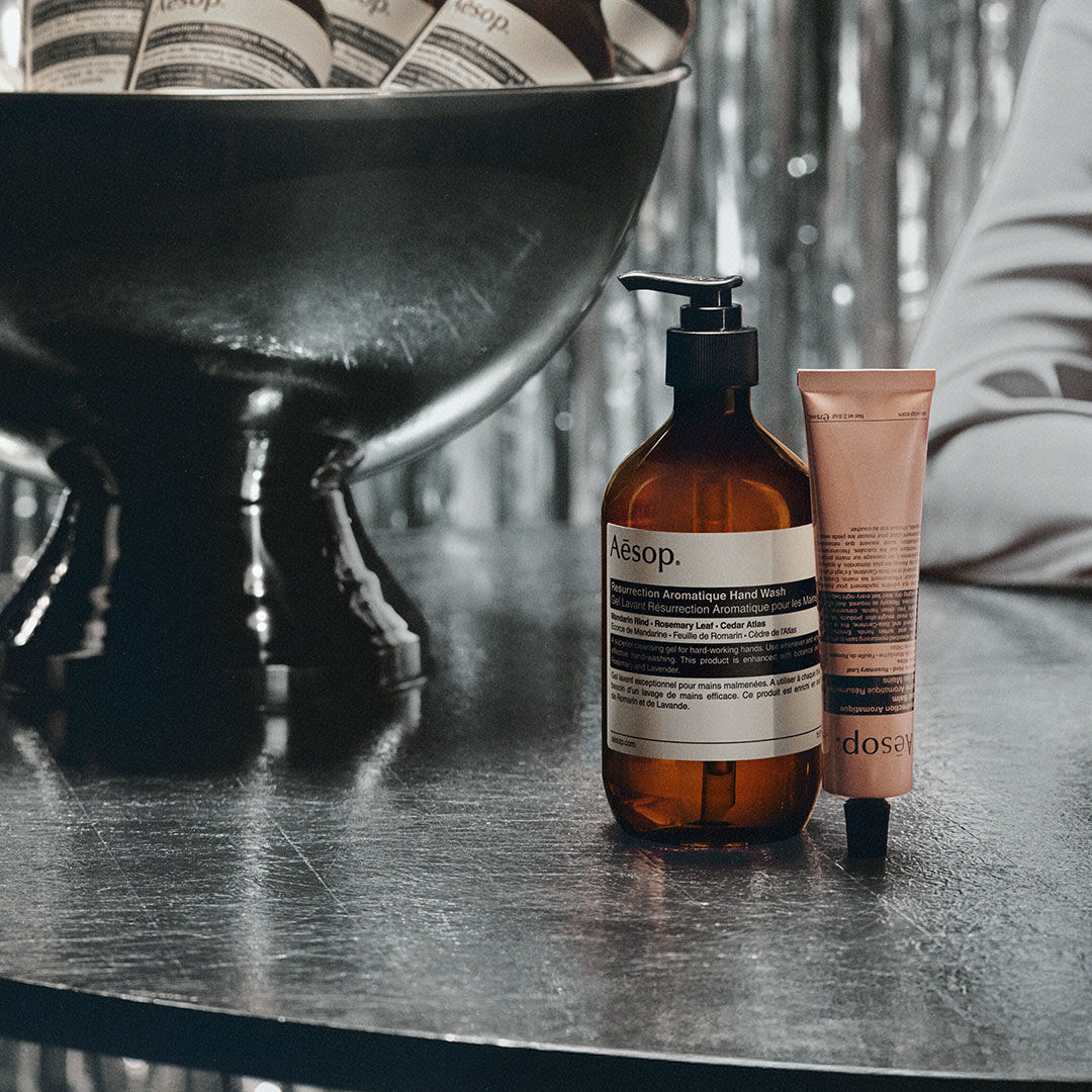 An Aesop hand wash bottle and hand cream tube beside a dark stone basin.