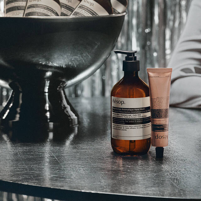 An Aesop hand wash bottle and hand cream tube beside a dark stone basin.
