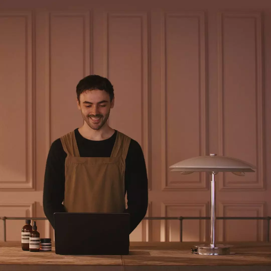 A smiling person in a brown apron looks at a laptop behind a wooden counter, beside amber bottles and a metal table lamp.
