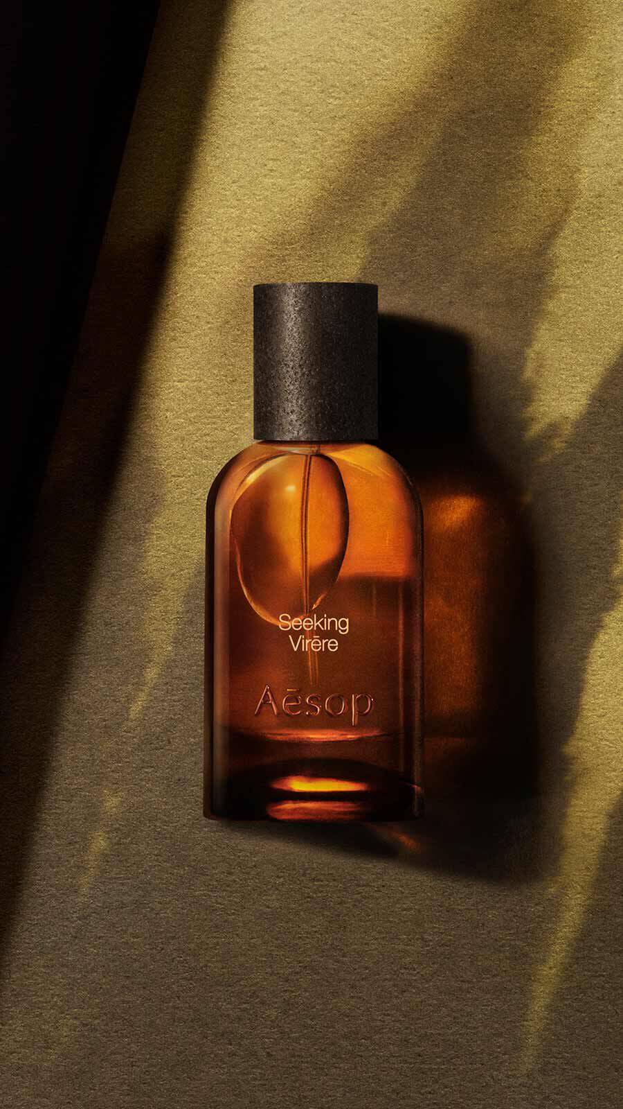 Aesop Seeking, Virere Eau de Parfum bottle with black cap on a warm golden-green surface with dramatic diagonal shadow lines.
