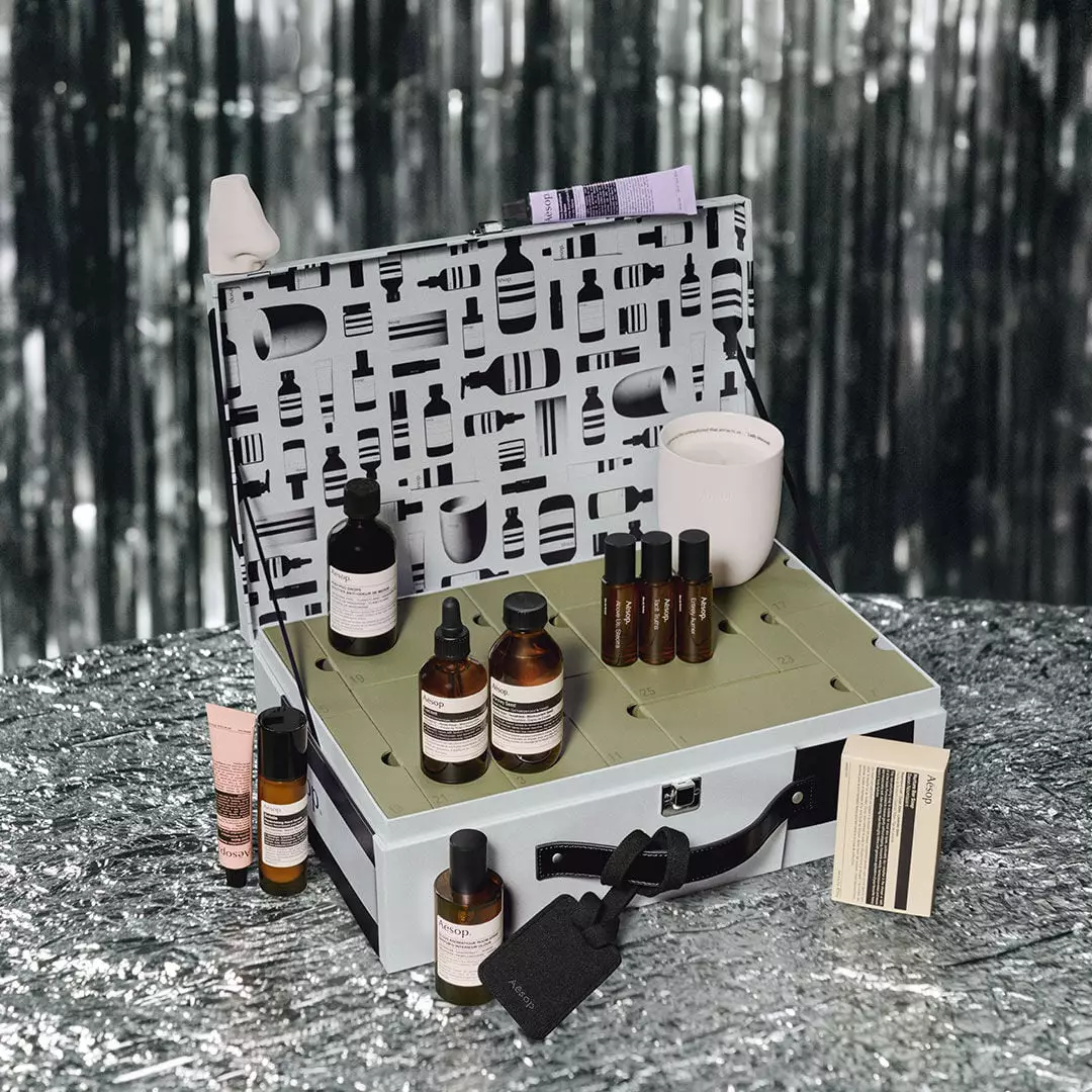 Aesop bottles, tubes and a white candle sit in and around an open silver case with numbered green compartments and a patterned lid.