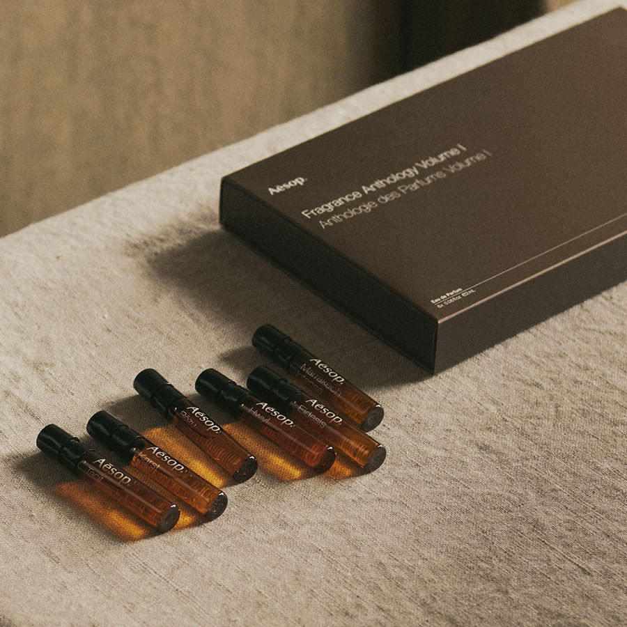 A collection of six small Aesop amber glass fragrance vials arranged on a linen surface next to their dark brown "Fragrance Anthology Volume I" presentation box.