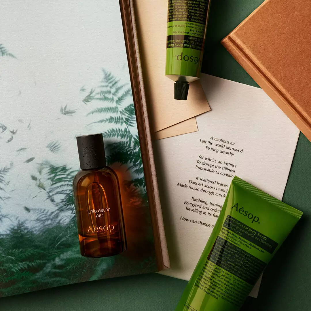 An Aesop Unforeseen Aer bottle rests on a fern print beside green skincare tubes, a printed poem and an orange book.