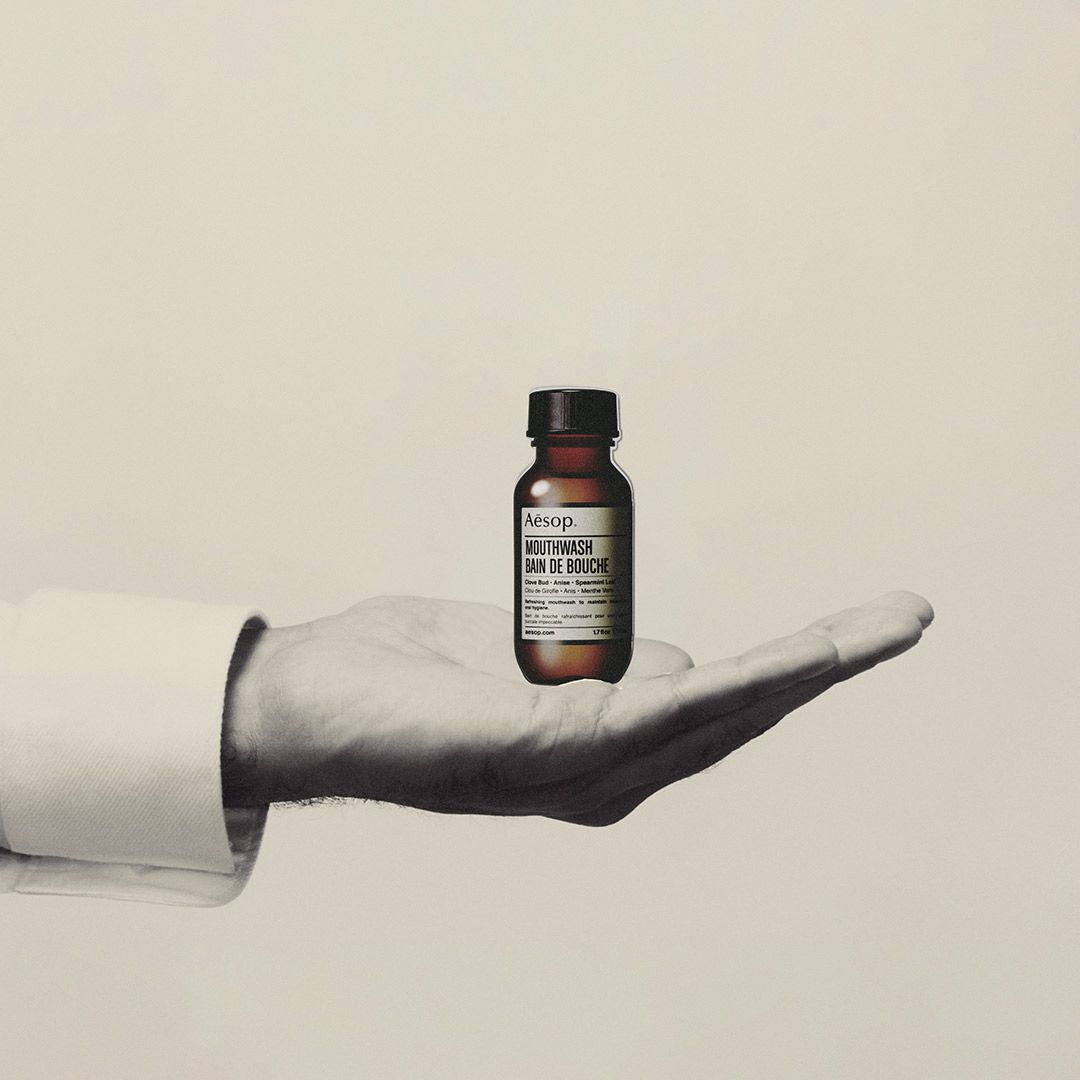 Hand holding a small amber Aesop mouthwash bottle against a plain background.