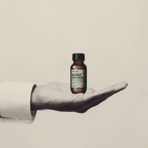 Hand holding a small amber Aesop mouthwash bottle against a plain background.