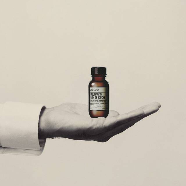 Hand holding a small amber Aesop mouthwash bottle against a plain background.