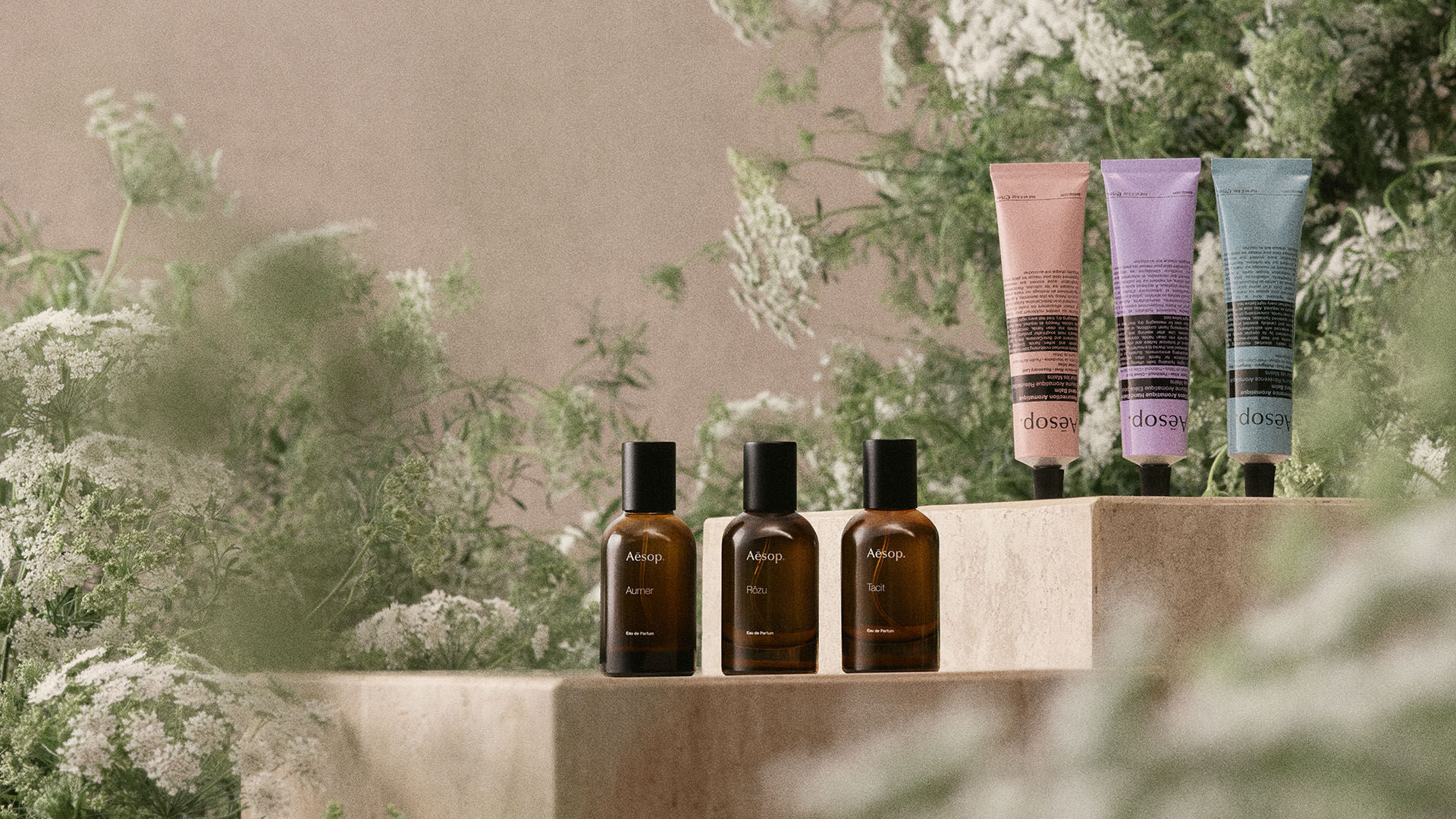 A still-life product photo featuring three Aesop amber glass perfume bottles and three colorful hand cream tubes arranged on stone pedestals amidst lush green foliage.
