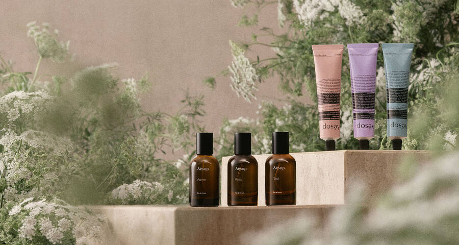 A still-life product photo featuring three Aesop amber glass perfume bottles and three colorful hand cream tubes arranged on stone pedestals amidst lush green foliage.