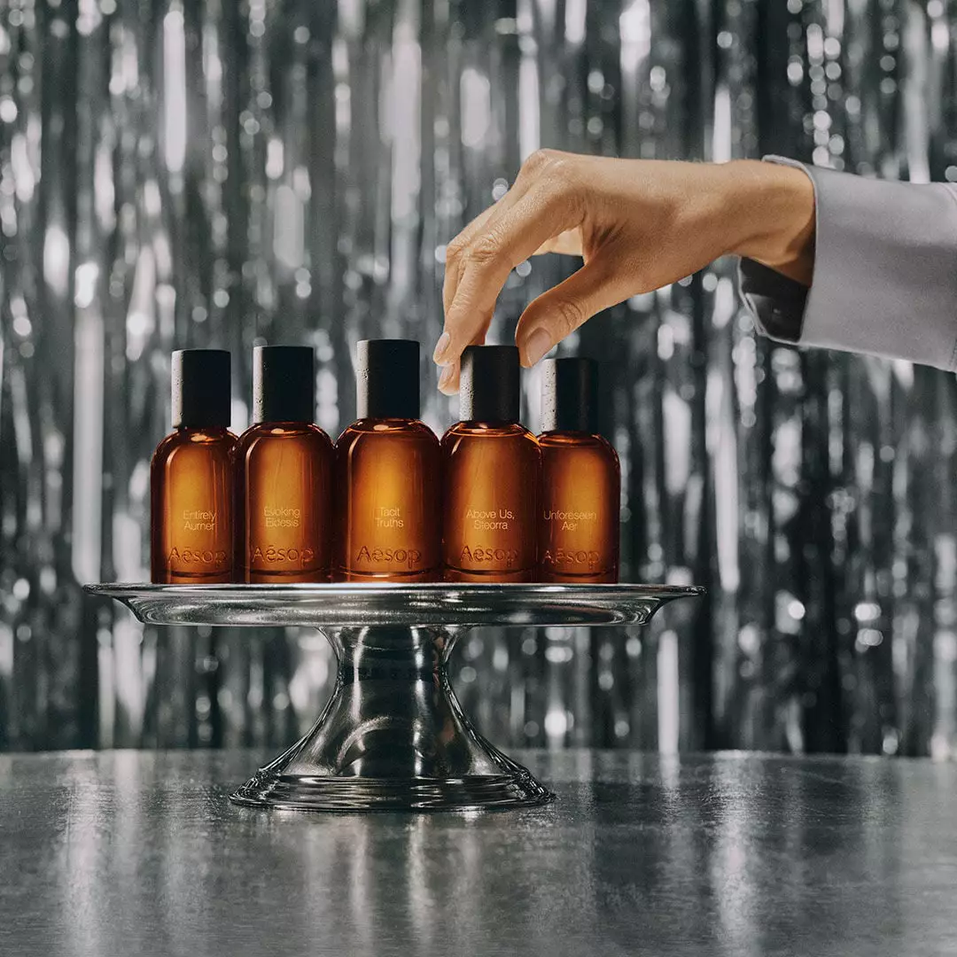 A hand touches the cap of one of five amber Aesop bottles arranged on a silver pedestal stand against silver tinsel.