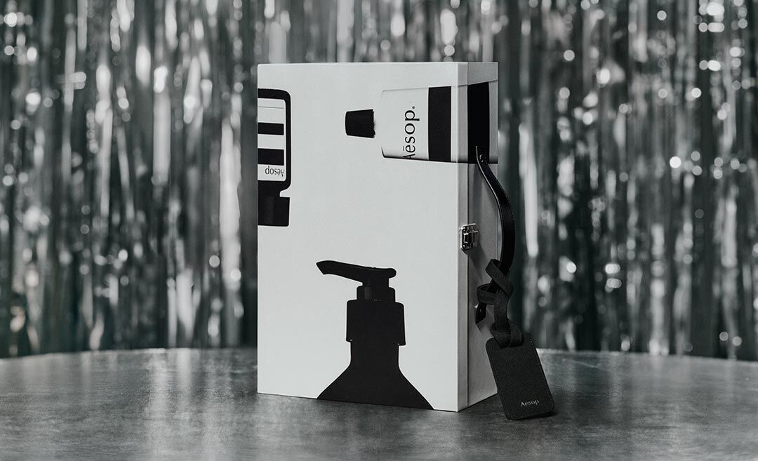 Upright silver Aesop case decorated with black bottle silhouettes, with a black luggage tag resting on a table against silver tinsel.
