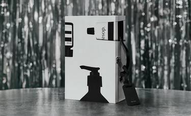Upright silver Aesop case decorated with black bottle silhouettes, with a black luggage tag resting on a table against silver tinsel.