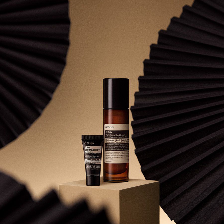 Aesop Solis hand serum bottle and tube displayed on a pedestal against a warm, minimalist backdrop.