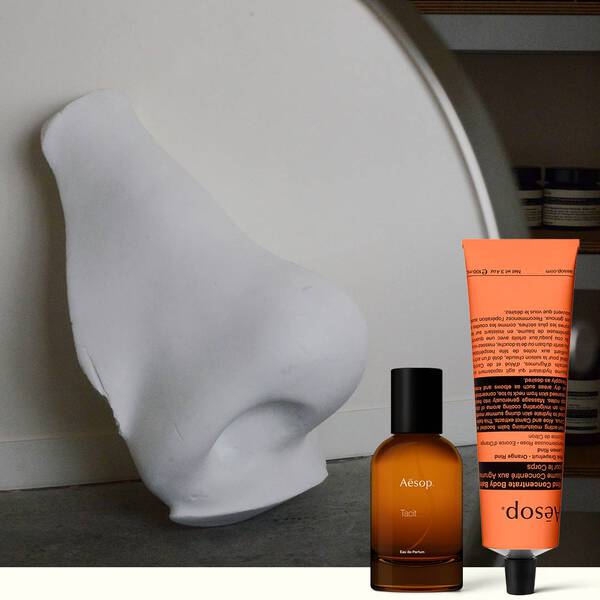White sculptural nose, amber Aesop perfume bottle, and orange Aesop tube on a dark background.