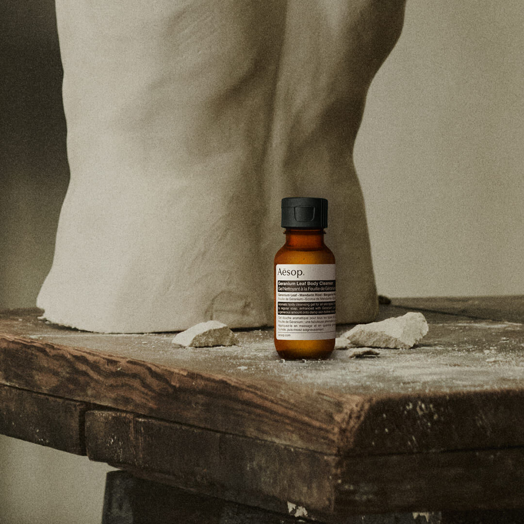 A small amber bottle of Aesop body cleanser on a rustic wooden table next to a clay torso sculpture.