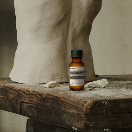 A small amber bottle of Aesop body cleanser on a rustic wooden table next to a clay torso sculpture.