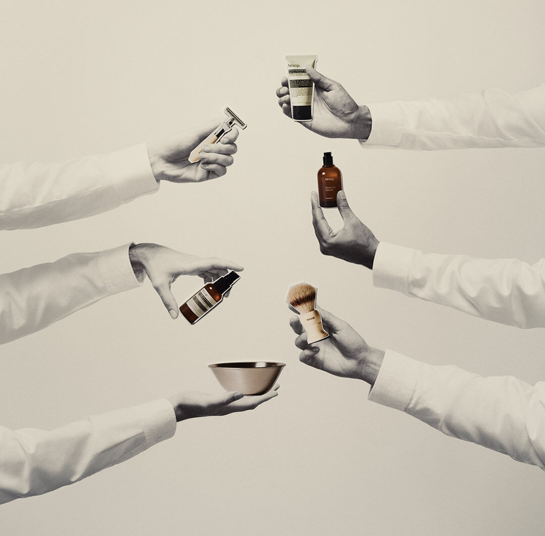 Four hands holding Aesop men's grooming products, including a safety razor, shaving serum, post-shave lotion, and fragrance, against a minimalist cream background.