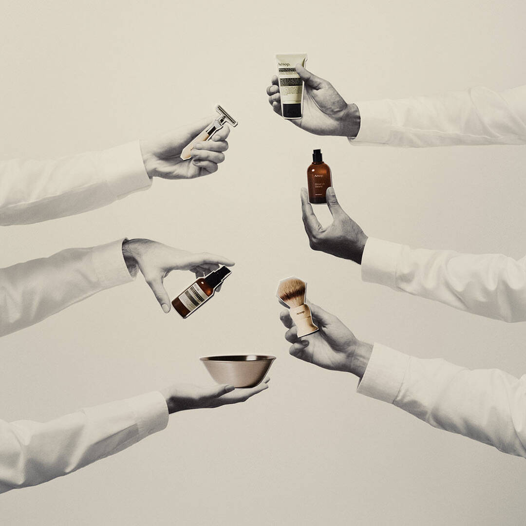 Four hands holding Aesop men's grooming products, including a safety razor, shaving serum, post-shave lotion, and fragrance, against a minimalist cream background.
