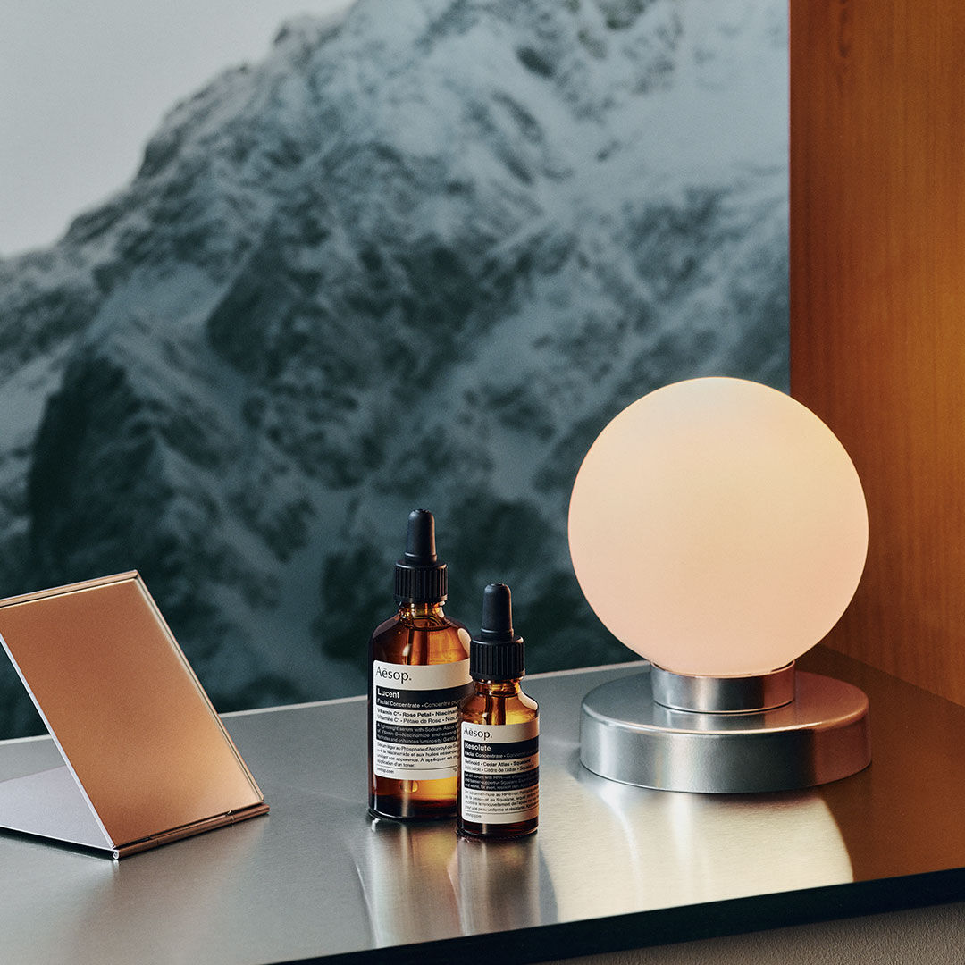 Two Aesop skincare bottles beside a glowing lamp, with snowy mountains in the background.
