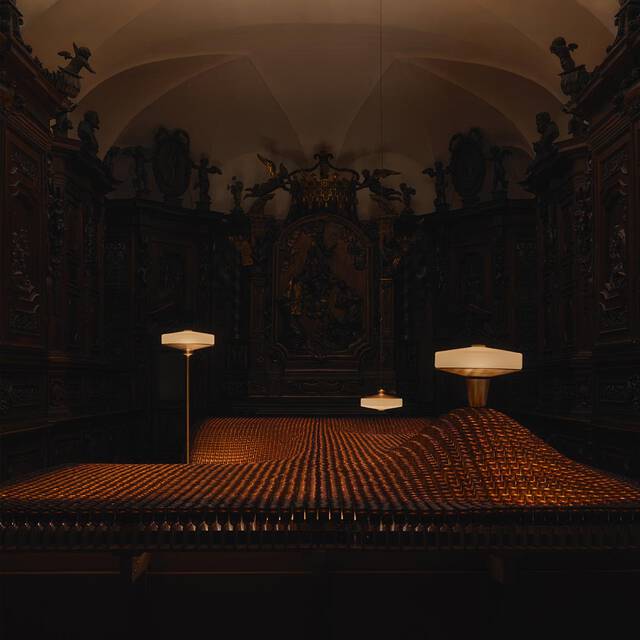 An artistic installation featuring thousands of amber glass bottles arranged in a glowing, wave-like landscape, set against a background of dark, ornate wood carvings and illuminated by minimalist modern lamps.
