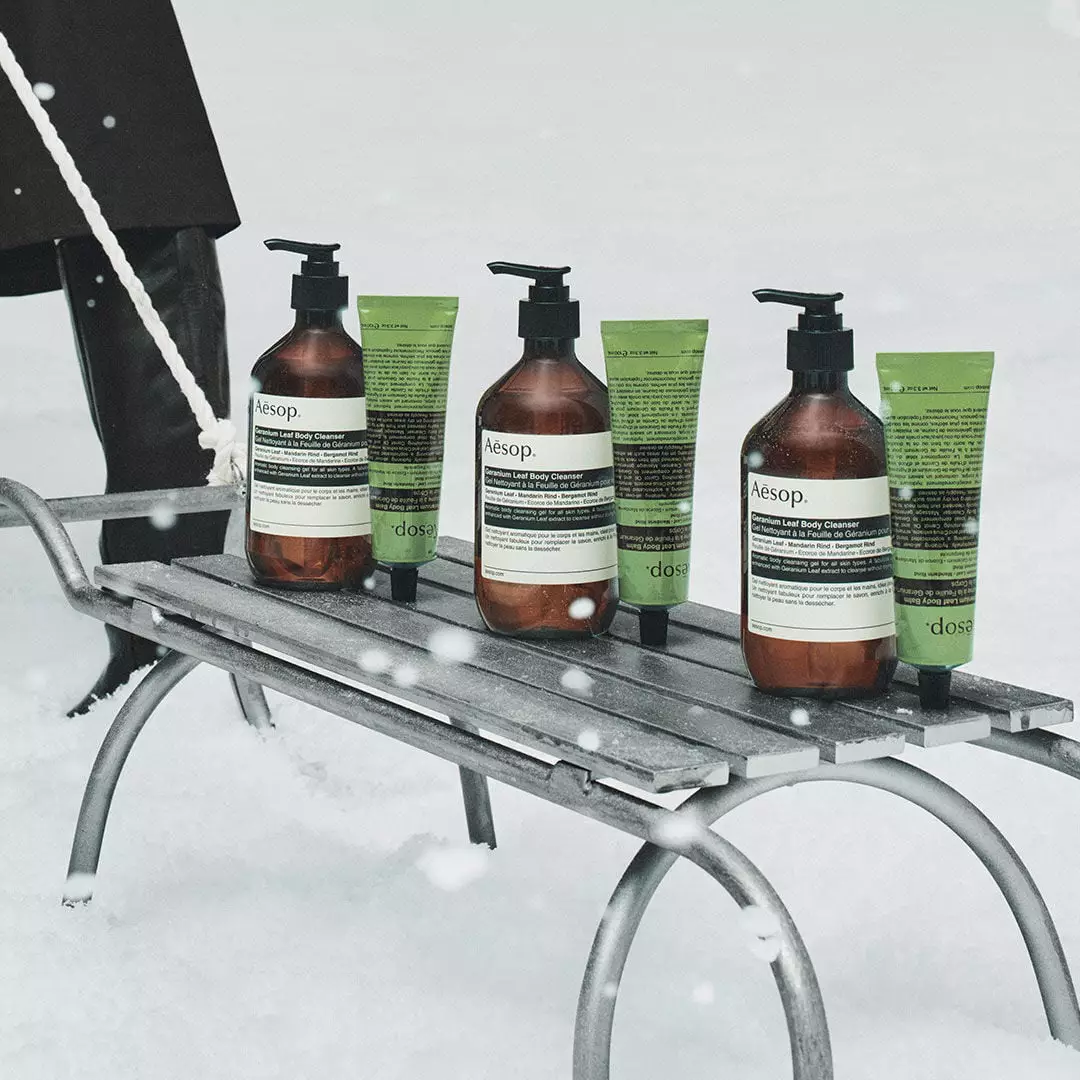 Three amber Aesop pump bottles alternate with three green tubes on a metal sledge in snow, beside a person's black boots.