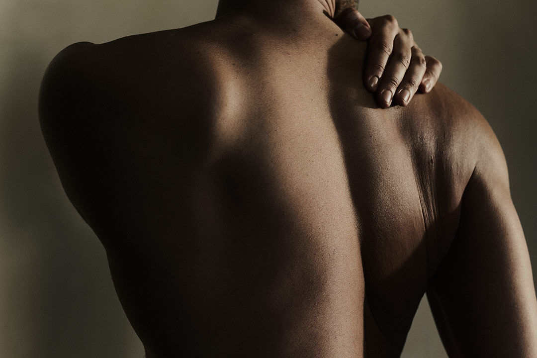 A close-up shot from behind of a person's bare upper back, with one hand resting on their shoulder under warm, dramatic lighting.